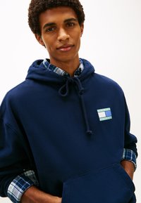 Μπλε ναυτικό φούτερ με κουκούλα και κορδόνι, με μικρό πολύχρωμο λογότυπο. Κάτω από αυτό, διακρίνεται ένα καρό πουκάμισο στους καρπούς.