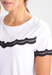 Mujer vestida con una camisa blanca de manga corta con encaje negro en el pecho y los bordes de las mangas, de pie contra un fondo claro y liso.