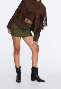 Bruine spijkerjack met franjes, olijfgroene rok met zakken en zwarte cowboylaarzen met een gestructureerd ontwerp en lage hak.