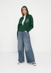 Zelena zip majica s velikim logotipom "GAP", uparena s širokim svijetloplavi trapericama i crnim tenisicama. Glatka tkanina i opušteni kroj.