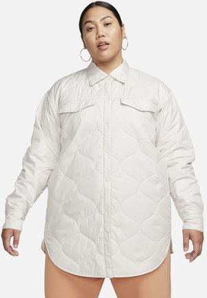 Γυναίκα φορώντας ένα λευκό μακριά μπουφάν με κουμπιά snap, τσέπες στο στήθος και σκουλαρίκια κρίκους, μπροστά σε ένα απλό φόντο.