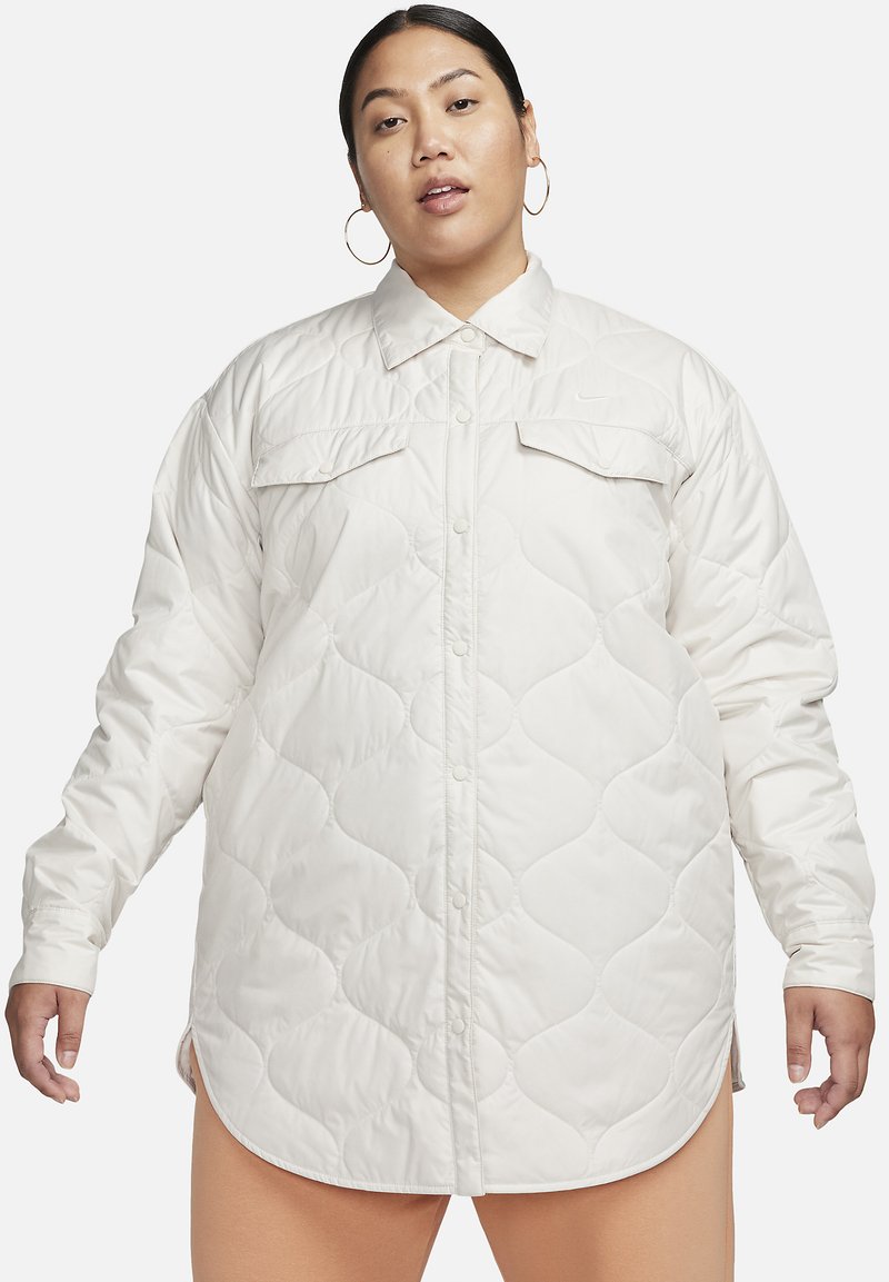 Femme portant une veste longue à manches longues en quilt blanc avec des boutons-pression, des poches poitrine et des boucles d'oreilles, sur un fond uni.