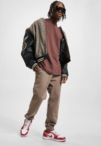 T-shirt marrón de gran tamaño, chaqueta bomber negra y de pata de gallo, joggers beige y zapatillas Nike rojas y blancas. Las gafas de sol completan el look.