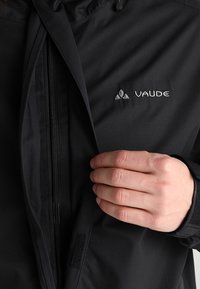 Black Vaude jacket with a zippered front, textured fabric, and embroidered logo. Features a snug fit with adjustable cuffs.