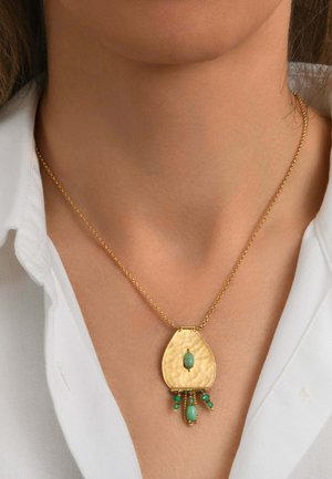 Collier en or avec un pendentif en forme de goutte martelée orné de pierres turquoises, porté sur une chemise blanche à col au cou d'une personne.