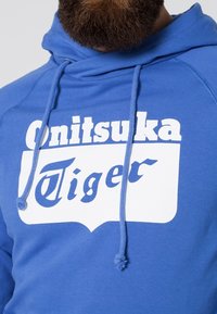 Sudadera azul con un logo blanco en el frente que dice "Onitsuka Tiger." Cuenta con cordones y una textura suave, con un diseño de ajuste holgado.