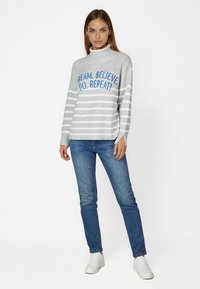 Gray striped sweater with turtleneck, featuring blue text: "DREAM. BELIEVE. DO. REPEAT!" Paired with blue jeans and white sneakers.