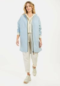 Lichtblauwe hoodie met rits en elastische manchetten, gedragen over een crèmekleurige top en beige broek, gecombineerd met beige sneakers.