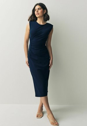 Form-fitting navy dress with shoulder pads and draped detailing. Smooth fabric, knee-length, paired with light, strappy sandals.