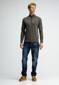 Grijze, gestructureerde pullover met een ritssluiting en een klein logo, gecombineerd met blauwe denim jeans en bruine suède schoenen, wat een casual outfit laat zien.