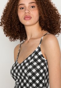 Top sem mangas em padrão xadrez preto e branco com um decote profundo e alças finas, feito de um tecido suave e texturizado. Pele levemente sardenta visível.