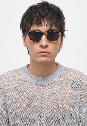 Pyöreät aurinkolasit, joissa on kilpikonnakuosiset kehykset ja tummat linssit. Mallilla on päällään vaaleanharmaa, neulottu villapaita, jossa on pyöreä pääntie.