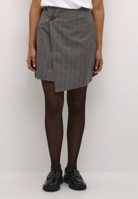 Jupe portefeuille en tartan gris avec une fermeture à bouton et une poche latérale, associée à des collants opaques noirs et des mocassins noirs brillants.