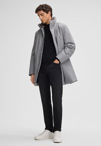 Grau melierter Wollmantel mit hohem Kragen, einem Knopfverschluss und seitlichen Taschen. Getragen über einem schwarzen Rollkragenpullover, kombiniert mit schwarzen Jeans und weißen Turnschuhen.