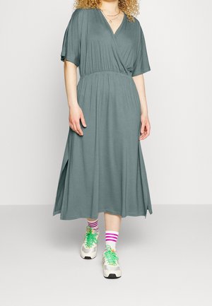 Vrouw draagt een grijze wikkeljurk tot halverwege de kuit met korte mouwen, witte sneakers met groene veters en witte sokken met roze strepen.