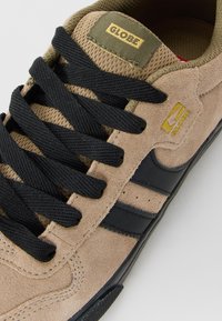 Tan semišové tenisky s černými tkaničkami a pruhy, s žlutým logem "Globe" na jazyku a na boku, a texturovanou olivově zelenou vnitřní podšívkou.