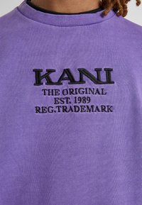 Felpa viola con testo ricamato nero che recita "KANI THE ORIGINAL EST. 1989 REG. TRADEMARK" sul davanti. Texture morbida.