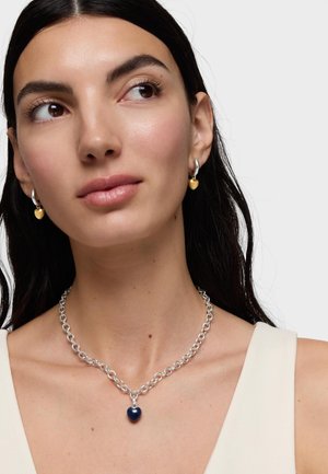 Mujer de cabello oscuro que lleva pendientes de aro plateados con charms en forma de corazón dorado y un collar grueso de cadena plateada con un colgante de corazón oscuro.