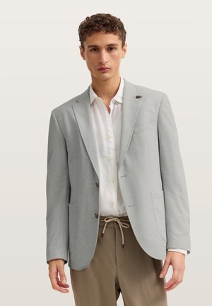 Junger Mann trägt hellgrauen Blazer, weißes Hemd mit Knöpfen und beige Hosen mit Kordelzug, steht vor schlichten Hintergrund.