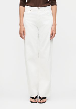 Pantaloni albi cu talie înaltă și croială largă, purtați cu sandale negre cu degetele descoperite și un top maro cu dungi, arătați de la talie până la picioare.