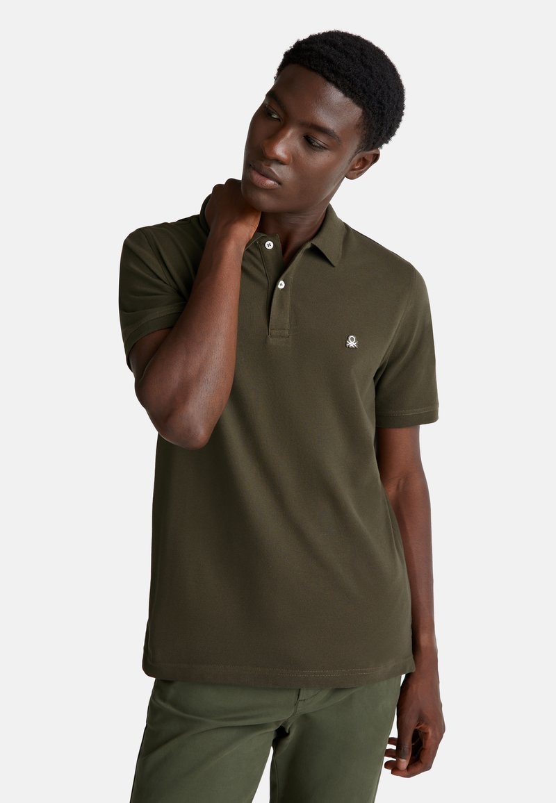 Muž v olivově zeleném polo tričku a kalhotách si sahá na krk, dívá se do strany na pozadí bílé stěny.