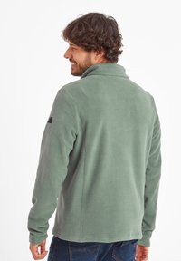 Fleece jacket in muted green, featuring a high collar and long sleeves. Smooth texture, rounded hem, and a small logo on the upper arm.