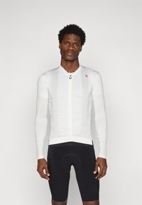Weißes Langarm-Radsporttrikot mit strukturiertem Design, Reißverschluss vorne und kleinem Logo. Trägt man über schwarzen Radsportshorts.