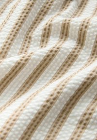 Witte stof met gestructureerde wafelweving en onregelmatige verticale beige strepen die een zacht, gebogen patroon vormen.