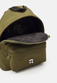 Zaino verde oliva con una fodera nera tessurizzata. Presenta una tasca frontale con zip, manico superiore e un logo sul davanti. Chiusura con zip.