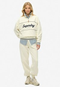 Cream-colored oversized sweatshirt with black logo, paired with cream sweatpants. Layered blue-striped shirt underneath. Worn with beige sneakers.