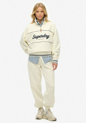 Kobieta stoi w kremowej bluzie Superdry, pod nią ma niebieską koszulę w paski, do tego pasujące kremowe dresy oraz beżowe sneakersy na białym tle.