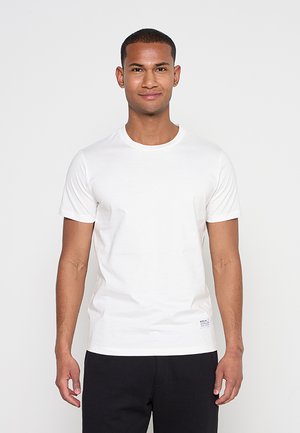 Junger Mann, der nach vorne schaut, trägt ein schlichtes weißes kurzärmeliges T-Shirt und schwarze Hose vor einem neutralen Hintergrund.