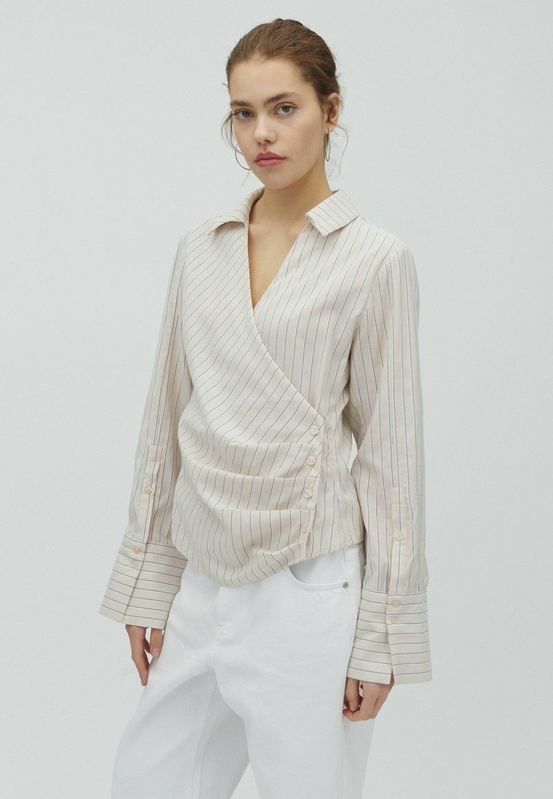 Ung kvinde iført en beige pinstripe slå-om-bluse med lange manchetter og hvide bukser mod en ensfarvet lys baggrund.