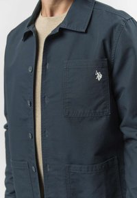 Tmavě námořnicky modrá košile z tkaniny s předním zapínáním na knoflíky, náprsní kapsou a malým logem. Má strukturovaný límec a rovný lem.