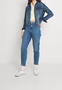 Persona con chaqueta de mezclilla azul y jeans, top corto amarillo claro, calcetines blancos y zapatillas altas blancas, de pie con una mano en la cadera.