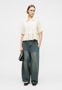 Γυναίκα που φοράει λευκό μπλουζάκι με κοντά μανίκια και πιέτες στη βάση, φαρδιά ξεβάμμένα μπλε τζιν παντελόνια, μαύρες μπαλαρίνες και κρατάει μία μαύρη τσάντα χειρός.