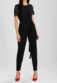 Zwarte jumpsuit met kanten korte mouwen, een ceintuur om de taille en taps toelopende benen, gecombineerd met rode hoge hakken tegen een neutrale achtergrond.
