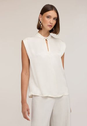 Mujer con cabello corto y castaño que lleva una blusa blanca sin mangas con pequeño escote en forma de ojo de cerradura y grandes pendientes de aro dorados.