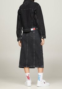 Personne portant une veste en denim noire et une jupe midi assortie, des baskets blanches et des chaussettes dépareillées avec du texte rouge et bleu.