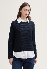 Pull en maille bleu marine avec un tissage texturé, col rond et manches longues, superposé à une chemise blanche à col, associé à un jean foncé.