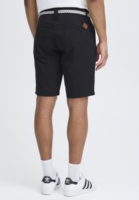 Shorts noirs avec une texture lisse, une ceinture avec un motif tissé blanc et une étiquette en cuir sur la taille. Baskets blanches visibles.