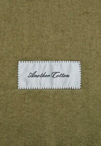 Tissu vert olive avec une texture douce ; présente une étiquette blanche cousue avec "Another Cotton" en lettres cursives noires.