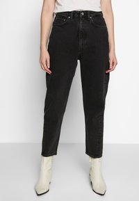 Jeans neri a vita alta realizzati in denim, con taglio dritto, cinque tasche e dettagli in bottone di rame, abbinati a stivaletti bianchi alla caviglia.