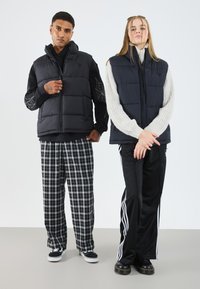 Gilet rembourré noir avec capuche et motif de toile d'araignée, porté par-dessus un sweat noir. Pull en tricot gris en dessous. Pantalons à carreaux avec des baskets noires.