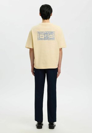 Osoba s krátkymi tmavými vlasmi, oblečená v béžovom tričku s modrým náčrtom výkladu obchodu a tmavých nohaviciach, stojaca zády k pozorovateľovi na jednoliatom pozadí.