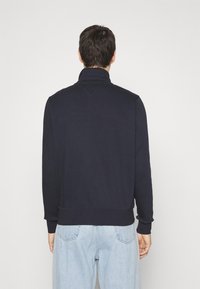 Persona di spalle che indossa una felpa nera con colletto alto e jeans azzurri contro uno sfondo bianco uniforme.
