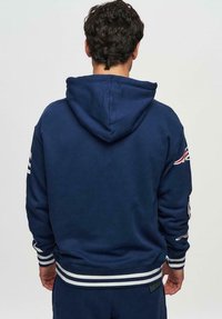 Mörkblå hoodie med en framficka i kängurustil, randiga muddar och fåll. Har en mönstrad axeldesign och justerbar huva.