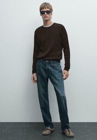 Giovane uomo che indossa un maglione a coste marrone scuro, jeans blu, scarpe mocassino beige e occhiali da sole neri, in piedi contro un muro chiaro semplice.