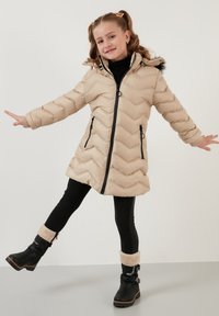 Beige pufferjakke med chevron-mønster, sort lynlås og lommer, hætte foret med pels, parret med sorte leggings og sorte støvler med pelskraver.