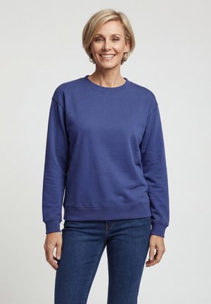 Lachende vrouw met kort blond haar, die een effen blauw sweatshirt met lange mouwen en blauwe spijkerbroek draagt, staand tegen een lichte achtergrond.